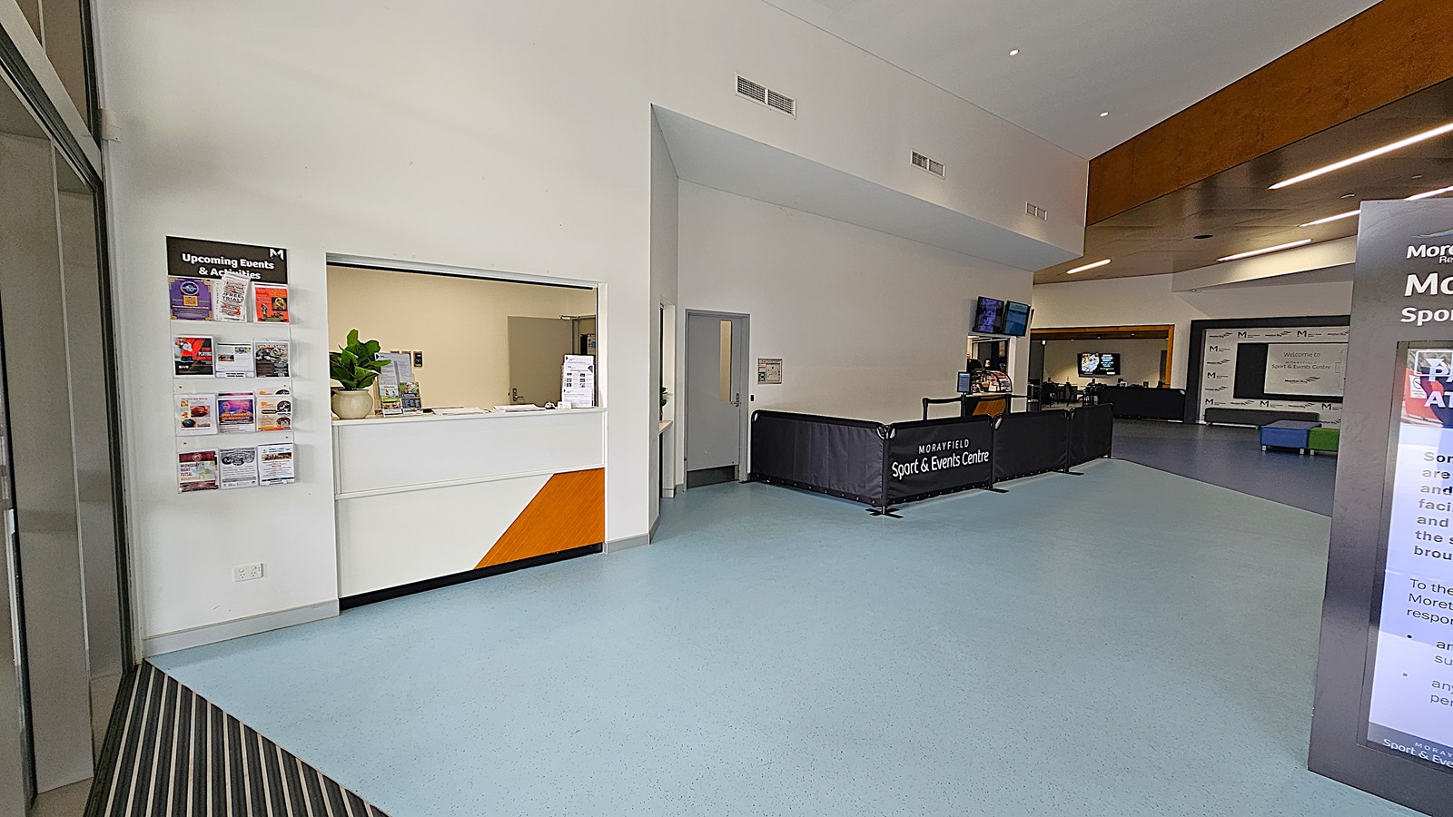 On the left, a large sign welcomes visitors to MSEC, with additional information displayed on nearby boards. The entrance features double glass doors that are open, revealing a spacious interior with a light blue floor. To the right, the entrance continues with another set of glass doors, and a dark counter is visible in the background, likely for reception. The ceiling is high and features recessed lighting. The ground is concrete, leading smoothly into the building.