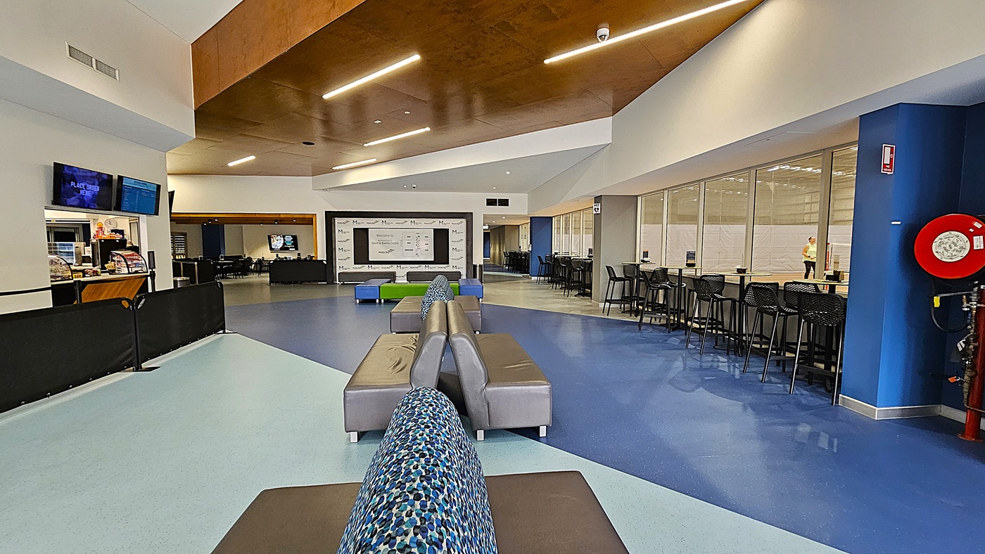 On the left, a counter area features a snack bar with various items displayed behind glass, alongside multiple screens showing information. A black barrier separates this area from the seating. The floor is a combination of light blue and dark blue tiles. In the centre, a grey sofa with a patterned backrest is positioned next to a green and blue seating arrangement. To the right, a corridor leads to additional rooms, with a few tables and chairs visible in the background.