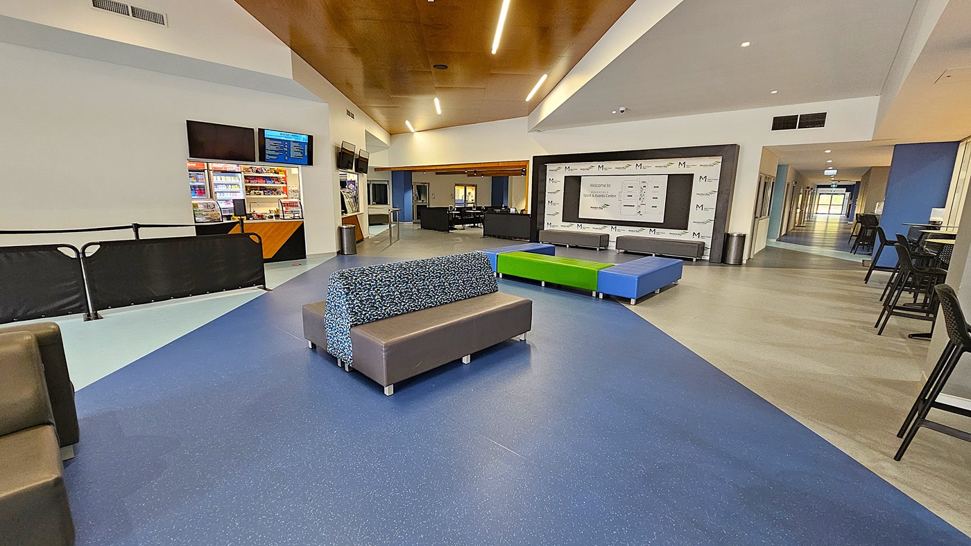 On the left, a reception area features a white counter with an orange accent, behind which is a plant and various informational materials displayed on a board. The floor is a light blue, smooth surface, leading towards the entrance. To the right, a black table with a banner for the "Moorfield Sport & Events Centre" is set up, alongside several screens displaying information. The ceiling is high and white, with recessed lighting.