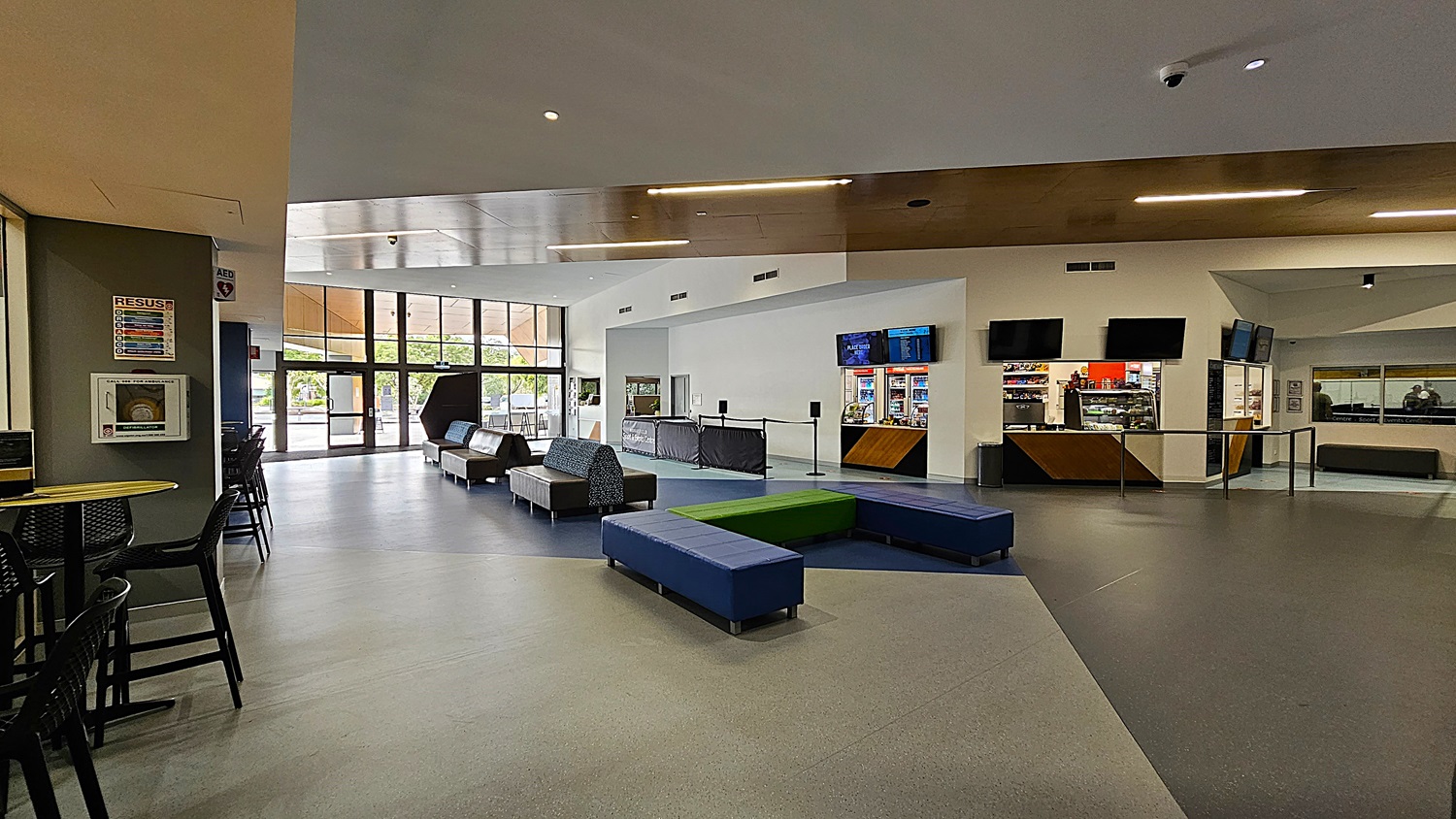 On the left, a small seating area features a round table with black chairs, alongside a wall-mounted first aid kit and a colourful poster. The floor is a combination of light grey and dark blue tiles, creating a geometric pattern. In the centre, a series of low benches in blue and green provide informal seating. To the right, a counter area with vending machines and screens displays information. The ceiling is a mix of white and wood, with recessed lighting.