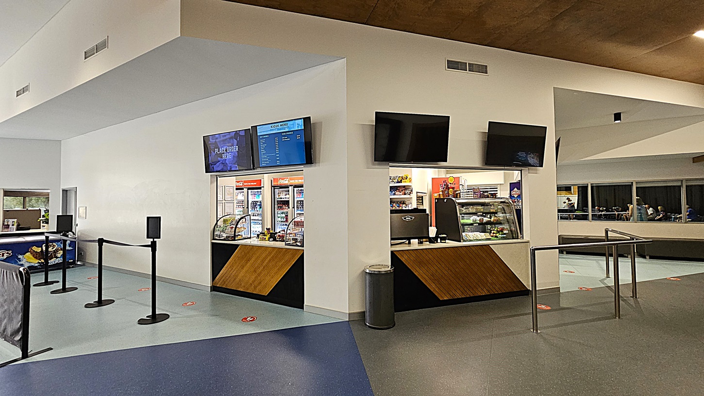 On the left, a counter area features a black barrier with stanchions for queue management, leading to a service point. The floor is a mix of light blue and dark blue tiles, creating a patterned effect. In the centre, a menu board is mounted above a glass display case filled with snacks and drinks. To the right, another counter is visible, equipped with a cash register and additional displays. The ceiling is smooth and light brown, with recessed lighting.