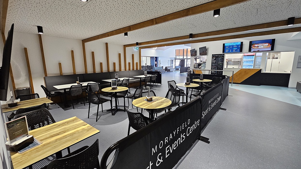 On the left, a series of round tables with wooden tops and black chairs are arranged, separated by a low black barrier displaying the name of the centre. The floor is a smooth grey surface, extending throughout the area. In the centre, a large open space leads to a counter with multiple screens displaying information and a menu. To the right, a service area features a glass display case and additional seating in the background. The ceiling is white with wooden beams.