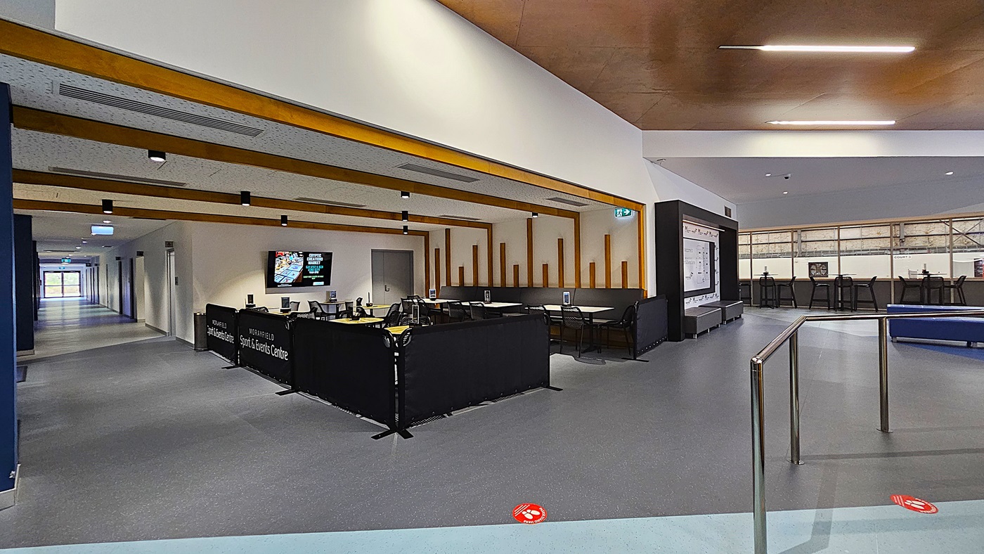 On the left, a reception counter is equipped with a black barrier and several screens displaying information. Behind the counter, there are seating areas with dark benches and tables. The floor is a smooth grey surface, extending throughout the space. In the centre, a corridor leads to other areas, with a light blue section visible. To the right, tall tables with black chairs are arranged against a wall, and a display board is mounted nearby. The ceiling features wooden beams and lighting.