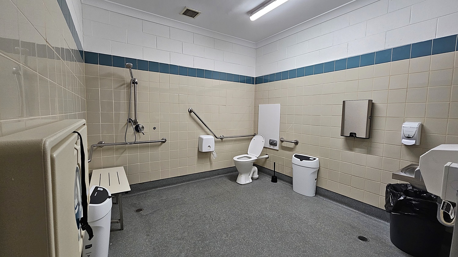 On the left, a beige wall features a toilet with grab rails beside it, and a small shelf for support. Below, a grey floor extends throughout the space. In the centre, a white toilet is positioned next to a black bin. To the right, a paper towel dispenser and a soap dispenser are mounted on the wall, along with a hand dryer. The walls are tiled in light beige with a blue accent strip at the top. The ceiling is white with fluorescent lighting.
