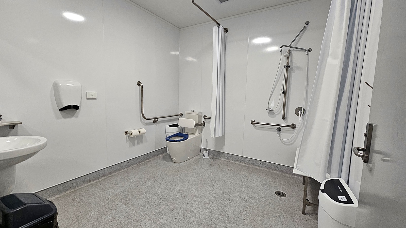 On the left, a white sink is mounted on the wall, with a black bin positioned beneath it. Above the sink, a soap dispenser is attached to the wall. The floor is a smooth grey surface, extending throughout the room. In the centre, a toilet with grab rails is visible, alongside a roll of toilet paper mounted on the wall. To the right, a shower area is enclosed by a curtain, with grab bars and a handheld showerhead.