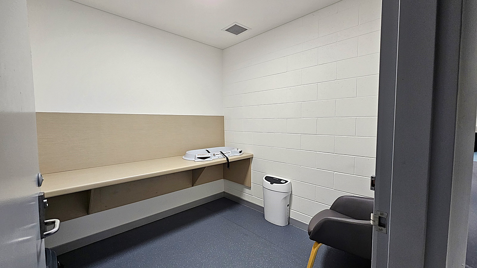 On the left, a long beige countertop runs along the wall, with a changing table positioned on top. A white bin is placed on the floor beside it. The floor is a dark blue, smooth surface. In the centre, the room is empty, providing ample space. To the right, a grey chair with wooden legs is positioned against the wall. The walls are painted white, with a beige panel running along the lower section. The ceiling is white with a ventilation grille.
