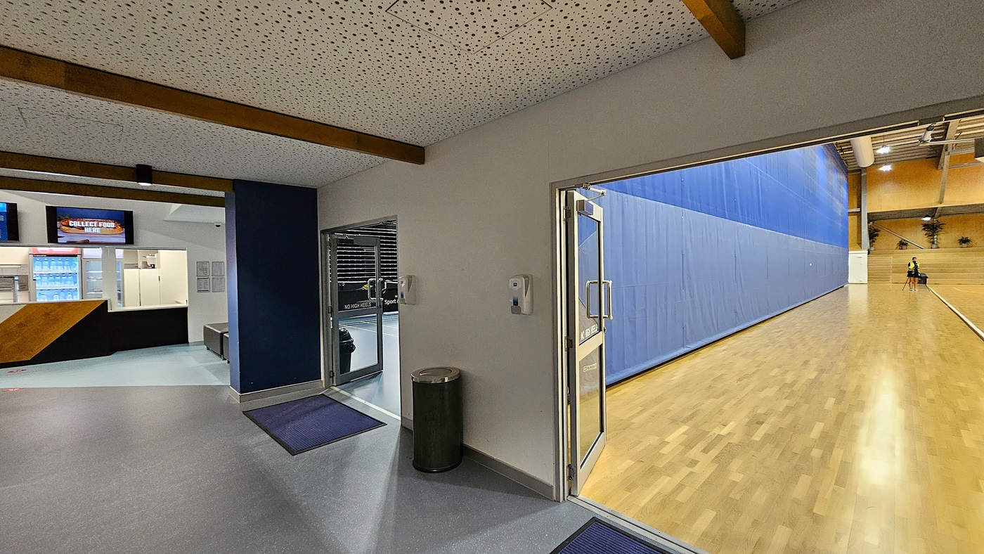 On the left, a grey door leads to a storage area, with a yellow wall beside it. The floor is a smooth blue surface marked with white lines for sports activities. In the centre, several table tennis tables are arranged, each with a net in place. To the right, a row of black chairs is lined up against the wall, next to a white table. The ceiling is high and features exposed beams with bright lights. In the background, tiered seating is visible against a black wall.