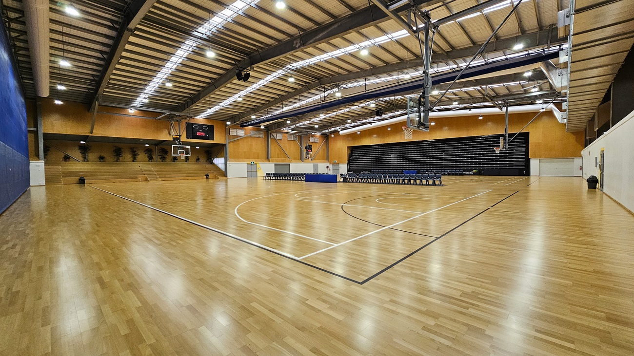 On the left, a blue wall features a door leading to a storage area, with tiered seating visible behind it. The floor is a polished wooden surface, marked with white lines for basketball. In the centre, a basketball hoop is mounted on the wall, with a digital scoreboard above displaying the time. To the right, a row of black chairs is arranged against a dark wall, with a closed door nearby. The ceiling is high, with exposed beams and bright lights.