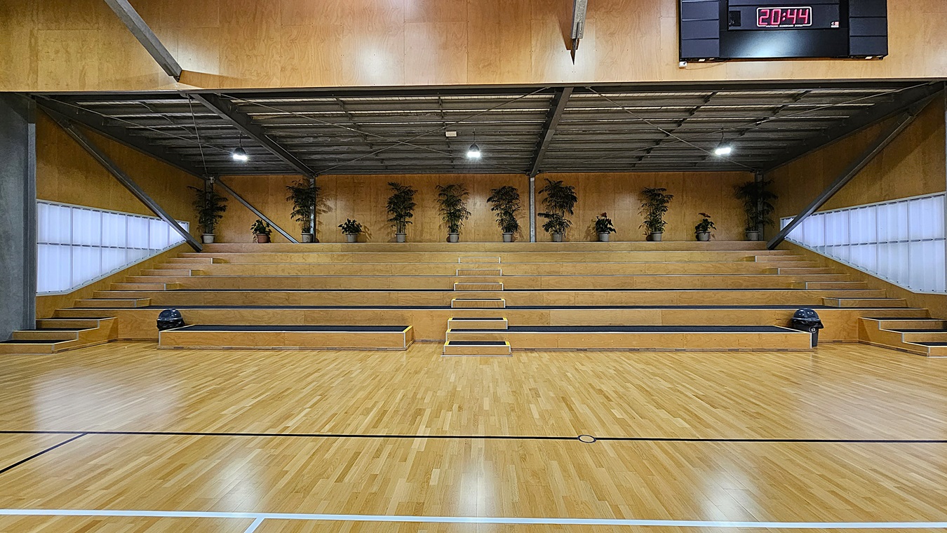 On the left, a set of steps leads up to a tiered seating area, which is lined with potted plants. The floor is a polished wooden surface, marked with black lines for sports activities. In the centre, the space is open, showcasing the wooden floor and the seating area in the background. To the right, additional steps lead to the upper tier of seating, with a scoreboard mounted on the wall displaying the time. The ceiling is high, featuring exposed beams and bright lights.