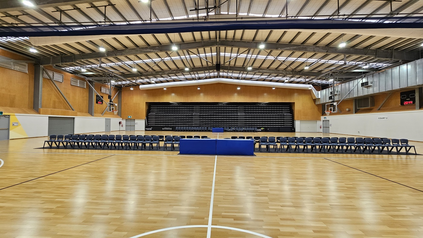 On the left, a row of blue chairs is arranged facing a central blue table, with a scoreboard visible above. The floor is a polished wooden surface, marked with white lines for basketball. In the centre, the court is open, leading to a tiered seating area in the background, which is filled with black seats. To the right, a basketball hoop is mounted on the wall, with a digital clock displaying the time. The ceiling is high, featuring exposed beams and bright lights.