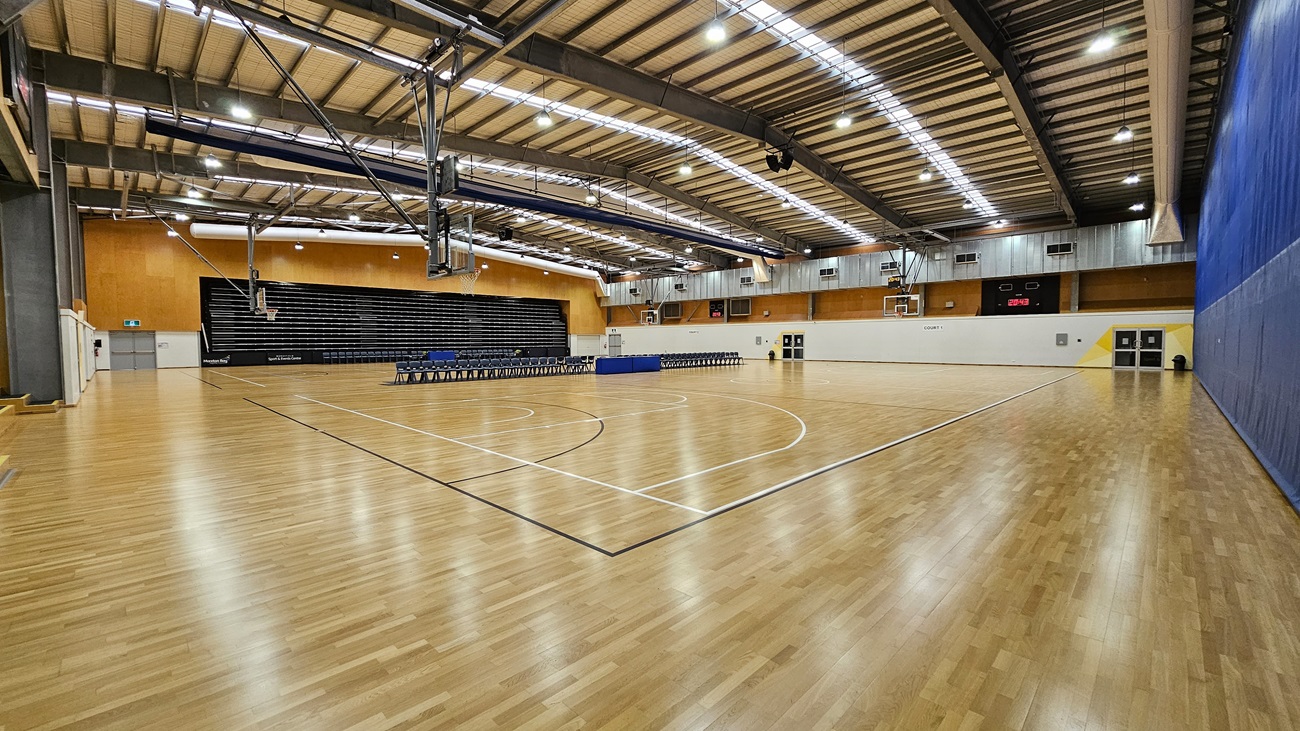 On the left, a set of steps leads to a tiered seating area, with black seats arranged against a wooden wall. The floor is a polished wooden surface, marked with white lines for basketball. In the centre, the court features a circular centre line and two basketball hoops mounted on the walls. To the right, a blue wall extends to the end of the court, with doors and a scoreboard visible. The ceiling is high, with exposed beams and bright lights.