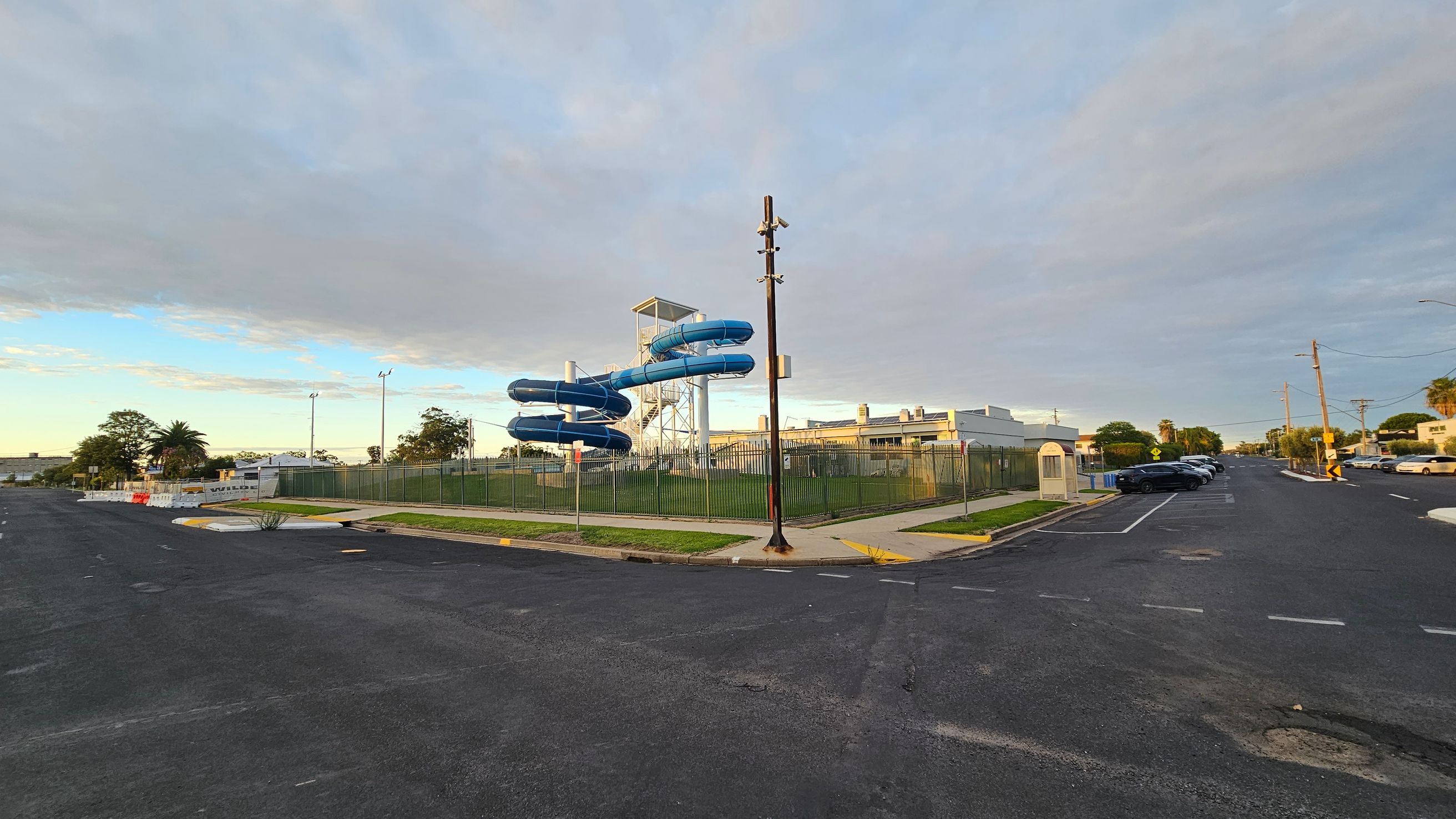 A street corner with a large blue water slide structure enclosed by a metal fence in the centre. The slide has multiple loops and a tall tower. On the left, there are trees and a construction barrier. On the right, a row of parked cars lines the street. A telephone pole stands near the corner, and a small white structure, possibly a phone booth, is visible. The pavement is dark, and the grass around the slide is green.
