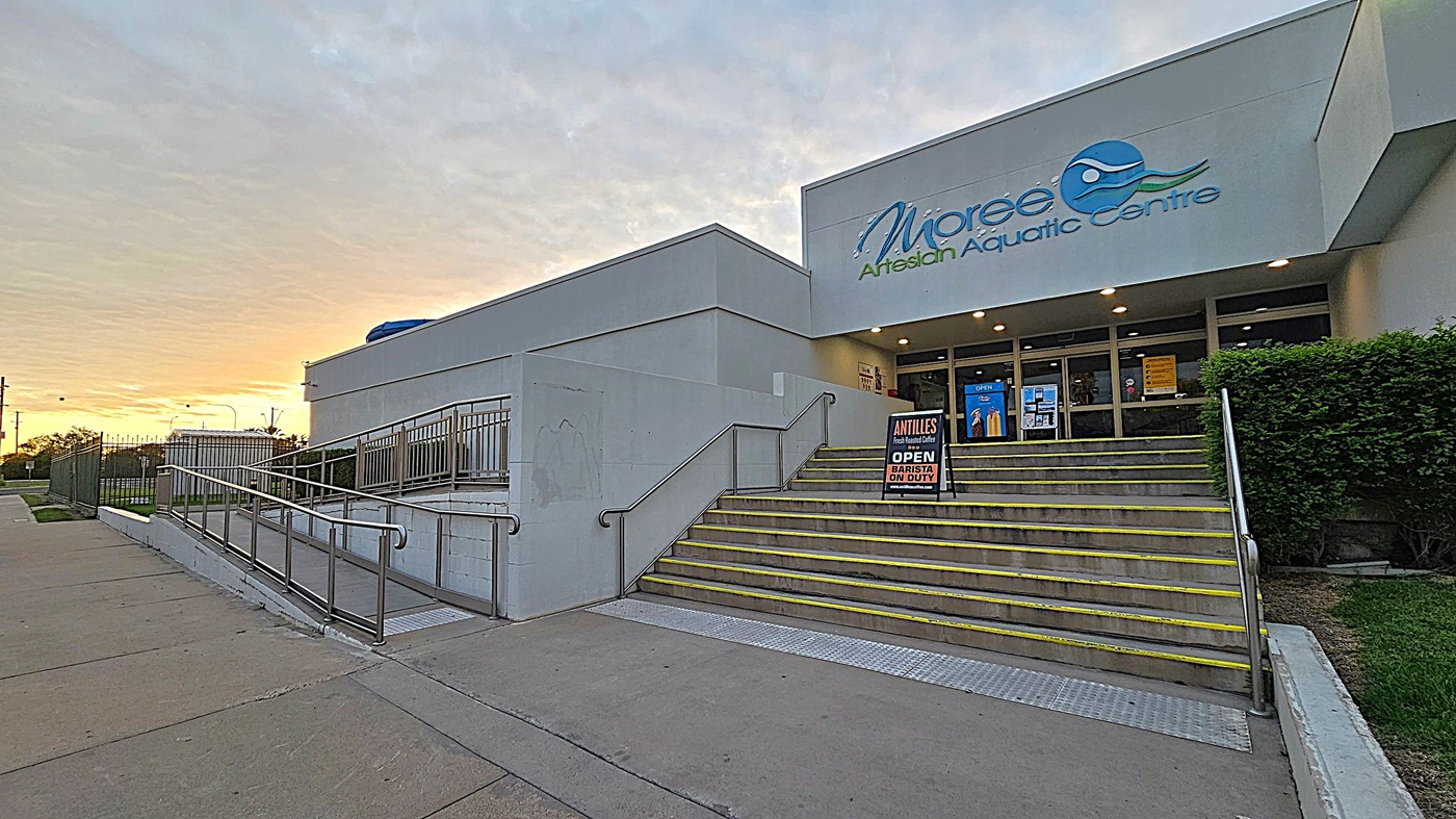 Entrance to a modern building with a sign reading "Moree Artesian Aquatic Centre" above the doors. On the right, a set of stairs with yellow-edged steps leads up to the entrance, accompanied by a sign advertising a café with a barista on duty. A metal railing runs alongside the stairs. On the left, a ramp with a metal railing provides an accessible route. The foreground features a concrete pavement, and there are bushes to the right of the entrance.