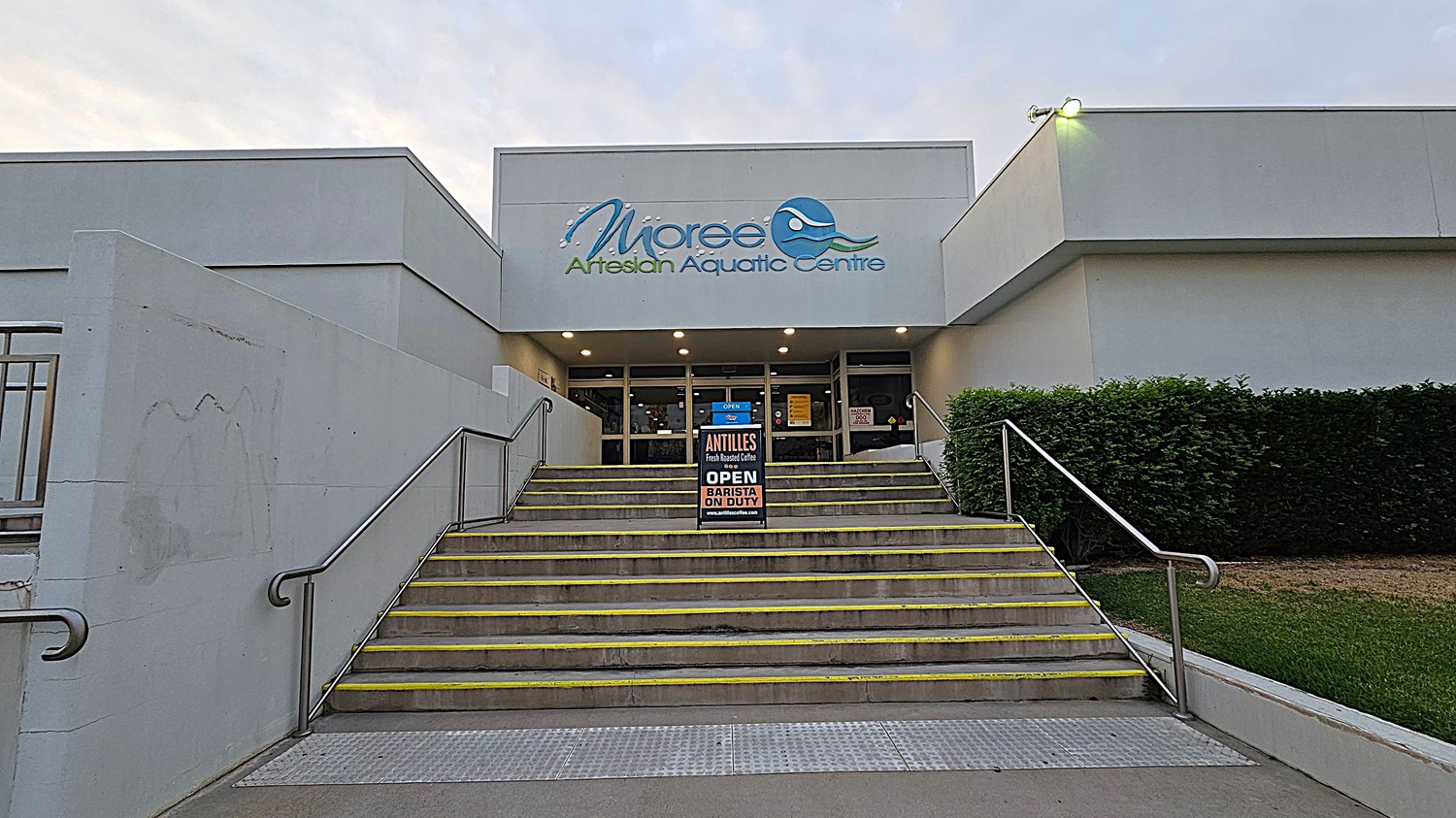 Entrance to a building with a sign reading "Moree Artesian Aquatic Centre" above the doors. A set of stairs with yellow-edged steps leads up to the entrance, flanked by metal railings. A sign on the steps advertises "Antilles Fresh Roasted Coffee" and mentions a barista on duty. On the right, there is a neatly trimmed hedge and grass. The building is modern with a grey facade and exterior lighting.