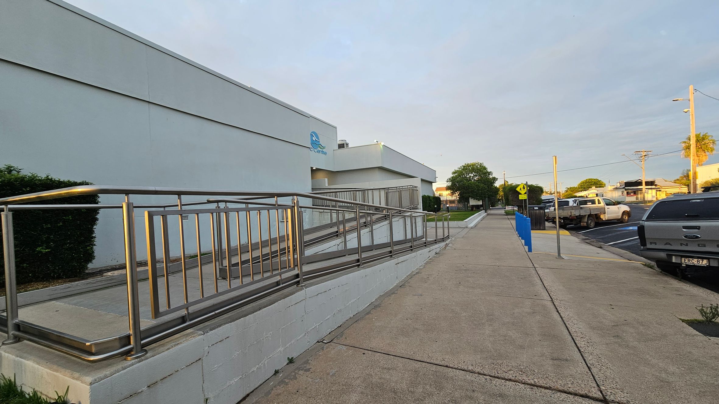 A building with a light blue exterior is on the left, featuring a metal handrail along a ramp. The pavement runs parallel to the building, leading to a parking area on the right. Several vehicles, including a grey truck, are parked along the street. A yellow pedestrian crossing sign is visible near the centre. Bushes and trees are scattered in the background, and a utility pole stands on the right.