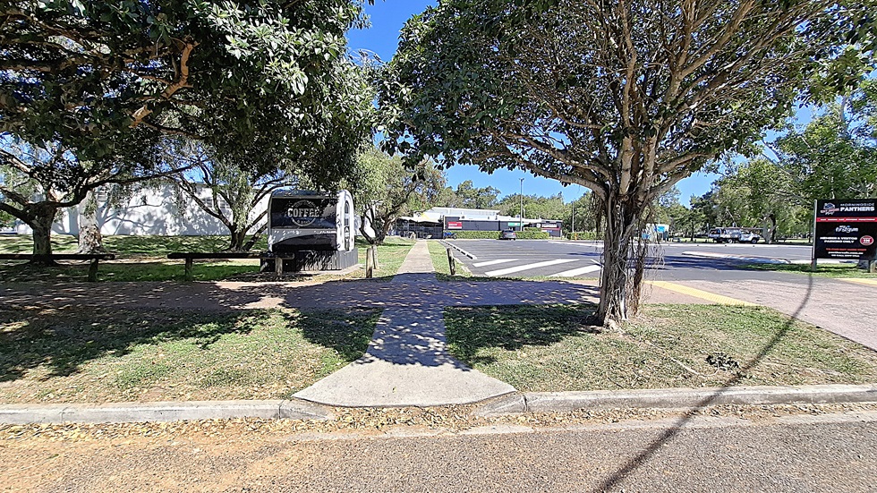 On the left, there are several large trees providing shade, with a grassy area visible behind them. A pathway leads through the greenery. To the right, a building with a white exterior is partially visible, along with a sign indicating "Panthers" and other businesses. The ground is a mix of asphalt and concrete, with a yellow tactile paving strip at the crossing. On the left, there are several large trees providing shade, with a grassy area visible behind them. A pathway leads through the greenery. To the right, a building with a white exterior is partially visible, along with a sign indicating "Panthers" and other businesses. The ground is a mix of asphalt and concrete, with a yellow tactile paving strip at the crossing.