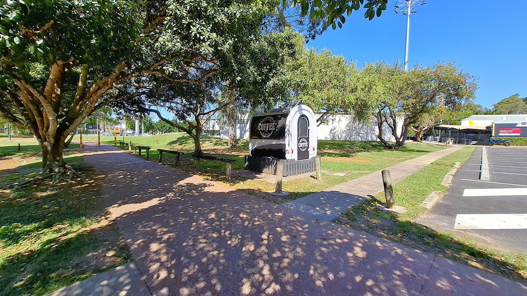 On the left, a black and white coffee truck is parked beside a path, surrounded by trees and grass. The path is made of compacted earth and is lined with benches. To the right, a concrete walkway leads towards a building with a white exterior, and further back, there is a parking area with marked spaces. The ground is a mix of grass and concrete, and the area appears well-maintained, with no visible litter. On the left, a black and white coffee truck is parked beside a path, surrounded by trees and grass. The path is made of compacted earth and is lined with benches. To the right, a concrete walkway leads towards a building with a white exterior, and further back, there is a parking area with marked spaces. The ground is a mix of grass and concrete, and the area appears well-maintained, with no visible litter.