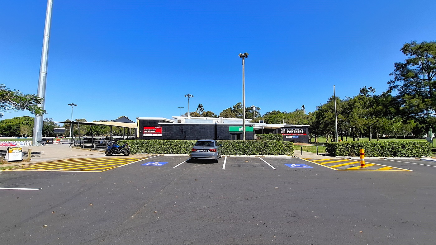 On the left, there is a parking area with a few marked spaces, including two designated for disabled parking, and a sign indicating the parking rules. A black motorcycle is parked nearby. To the right, a building with a black exterior features signs for "Panthers" and other businesses. The ground is asphalt, clearly marked with yellow lines. In the background, trees provide greenery, and there are light poles visible. On the left, there is a parking area with a few marked spaces, including two designated for disabled parking, and a sign indicating the parking rules. A black motorcycle is parked nearby. To the right, a building with a black exterior features signs for "Panthers" and other businesses. The ground is asphalt, clearly marked with yellow lines. In the background, trees provide greenery, and there are light poles visible.