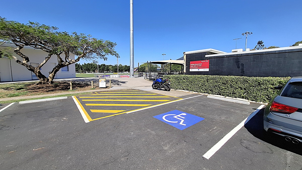 On the left, a large tree provides shade over a grassy area, with a few wooden posts visible nearby. A parking space is marked with yellow lines, including a blue symbol for disabled parking. A blue motorcycle is parked in this space. To the right, a building with a black exterior features a sign indicating "Athletics Queensland." The ground is asphalt, clearly marked, and the area appears tidy, with a low hedge bordering the parking lot. On the left, a large tree provides shade over a grassy area, with a few wooden posts visible nearby. A parking space is marked with yellow lines, including a blue symbol for disabled parking. A blue motorcycle is parked in this space. To the right, a building with a black exterior features a sign indicating "Athletics Queensland." The ground is asphalt, clearly marked, and the area appears tidy, with a low hedge bordering the parking lot.