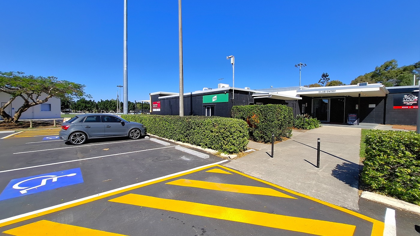 On the left, a grey car is parked in a designated disabled parking space, marked with a blue symbol. A tree with green foliage is visible behind the car, providing some shade. To the right, a building with a black exterior features a green sign indicating "Club Entry." The entrance is flanked by neatly trimmed hedges. The ground is asphalt with yellow diagonal lines marking the parking area. On the left, a grey car is parked in a designated disabled parking space, marked with a blue symbol. A tree with green foliage is visible behind the car, providing some shade. To the right, a building with a black exterior features a green sign indicating "Club Entry." The entrance is flanked by neatly trimmed hedges. The ground is asphalt with yellow diagonal lines marking the parking area.