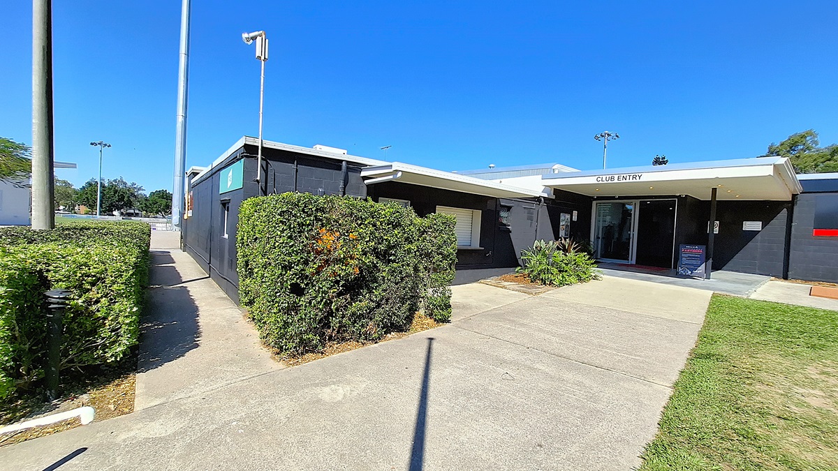 On the left, a pathway bordered by neatly trimmed hedges leads towards a building. The path is made of concrete and is well-maintained. To the right, the building features a black exterior with a sign reading "CLUB ENTRY" above the entrance. The entrance is framed by a small overhang, and there are potted plants beside the door. The ground is also concrete, with a slight incline towards the entrance. On the left, a pathway bordered by neatly trimmed hedges leads towards a building. The path is made of concrete and is well-maintained. To the right, the building features a black exterior with a sign reading "CLUB ENTRY" above the entrance. The entrance is framed by a small overhang, and there are potted plants beside the door. The ground is also concrete, with a slight incline towards the entrance.