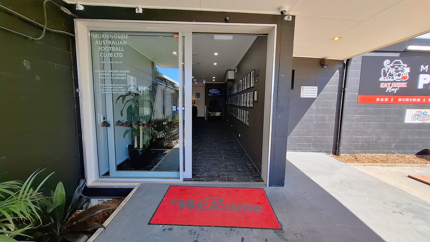 On the left, a glass door leads into a corridor with a tiled floor, and a few potted plants are positioned near the entrance. A red welcome mat is placed at the threshold. To the right, a wall displays several framed photographs, and a small air conditioning unit is mounted above. The ceiling features recessed lighting, providing illumination to the area. The floor is dark tiled, extending into the corridor, which appears clean and well-maintained. On the left, a glass door leads into a corridor with a tiled floor, and a few potted plants are positioned near the entrance. A red welcome mat is placed at the threshold. To the right, a wall displays several framed photographs, and a small air conditioning unit is mounted above. The ceiling features recessed lighting, providing illumination to the area. The floor is dark tiled, extending into the corridor, which appears clean and well-maintained.