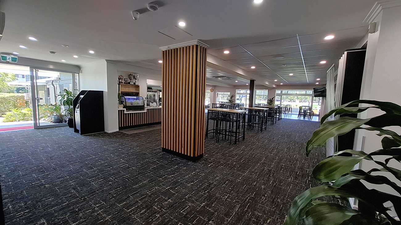 On the left, a glass door opens to the outside, with a small area featuring potted plants. Adjacent to the door, there are two black machines, possibly for ordering or payment. In the centre, a wooden bar counter with a glass display case is visible, showcasing items for sale. To the right, tall tables with black stools are arranged, and large windows allow light into the space. The floor is covered in dark carpet with a textured pattern, and the ceiling features recessed lighting. On the left, a glass door opens to the outside, with a small area featuring potted plants. Adjacent to the door, there are two black machines, possibly for ordering or payment. In the centre, a wooden bar counter with a glass display case is visible, showcasing items for sale. To the right, tall tables with black stools are arranged, and large windows allow light into the space. The floor is covered in dark carpet with a textured pattern, and the ceiling features recessed lighting.