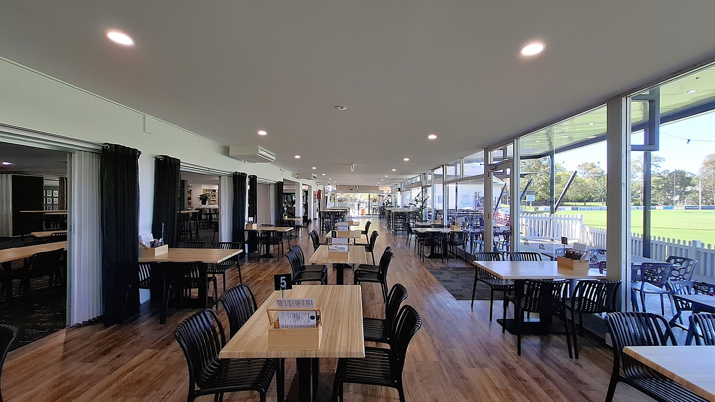On the left, a series of black curtains frame an entrance, leading to a more private area. Several wooden tables with black chairs are arranged in the foreground, with a small menu holder on one table. In the centre, the floor is made of polished wooden boards, creating a warm appearance. To the right, large glass windows allow light to flood the space, revealing outdoor seating and greenery beyond. The ceiling is white with recessed lighting. On the left, a series of black curtains frame an entrance, leading to a more private area. Several wooden tables with black chairs are arranged in the foreground, with a small menu holder on one table. In the centre, the floor is made of polished wooden boards, creating a warm appearance. To the right, large glass windows allow light to flood the space, revealing outdoor seating and greenery beyond. The ceiling is white with recessed lighting.