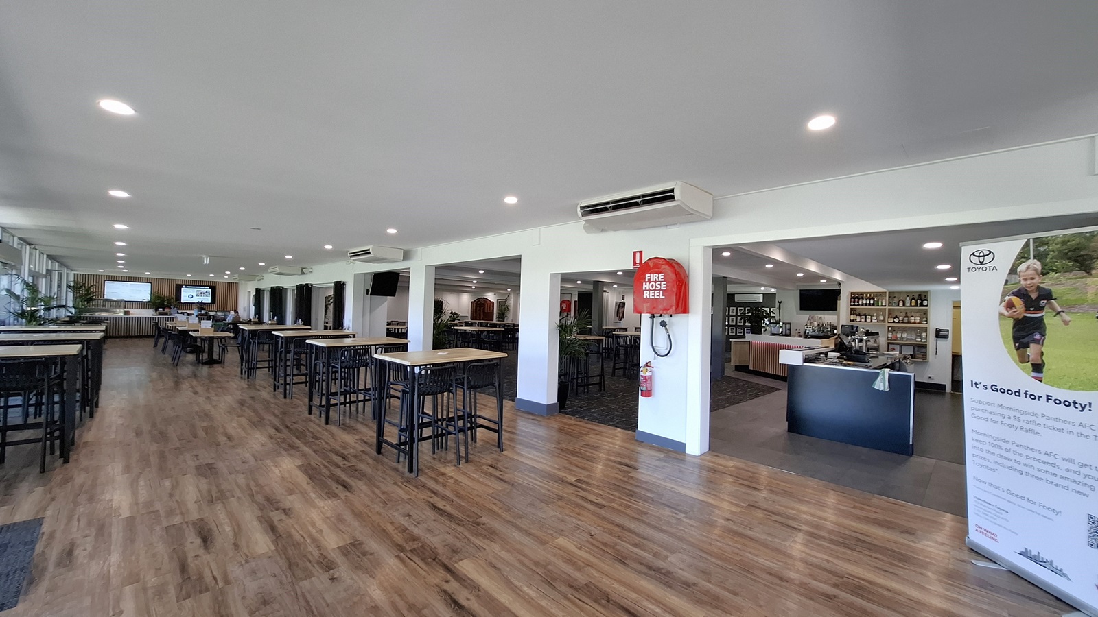 On the left, several tall tables with black stools are arranged on a polished wooden floor, creating a spacious dining area. A large window allows natural light to enter, with potted plants positioned nearby. In the centre, the floor continues with the same wooden finish, leading towards a bar area. To the right, a wall features a large banner promoting a local event, alongside a black counter with bottles displayed behind it. The ceiling is white with recessed lighting. On the left, several tall tables with black stools are arranged on a polished wooden floor, creating a spacious dining area. A large window allows natural light to enter, with potted plants positioned nearby. In the centre, the floor continues with the same wooden finish, leading towards a bar area. To the right, a wall features a large banner promoting a local event, alongside a black counter with bottles displayed behind it. The ceiling is white with recessed lighting.