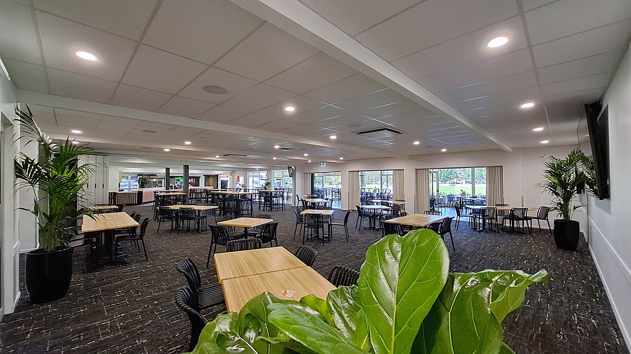 On the left, a large potted plant adds greenery beside a white wall. Several wooden tables with black chairs are arranged across the dark carpeted floor, creating a dining area. In the centre, the space is open, with more tables visible, and a bar counter can be seen in the background. To the right, large glass doors lead outside, framed by light curtains, allowing natural light to fill the room. The ceiling is white with recessed lighting. On the left, a large potted plant adds greenery beside a white wall. Several wooden tables with black chairs are arranged across the dark carpeted floor, creating a dining area. In the centre, the space is open, with more tables visible, and a bar counter can be seen in the background. To the right, large glass doors lead outside, framed by light curtains, allowing natural light to fill the room. The ceiling is white with recessed lighting.