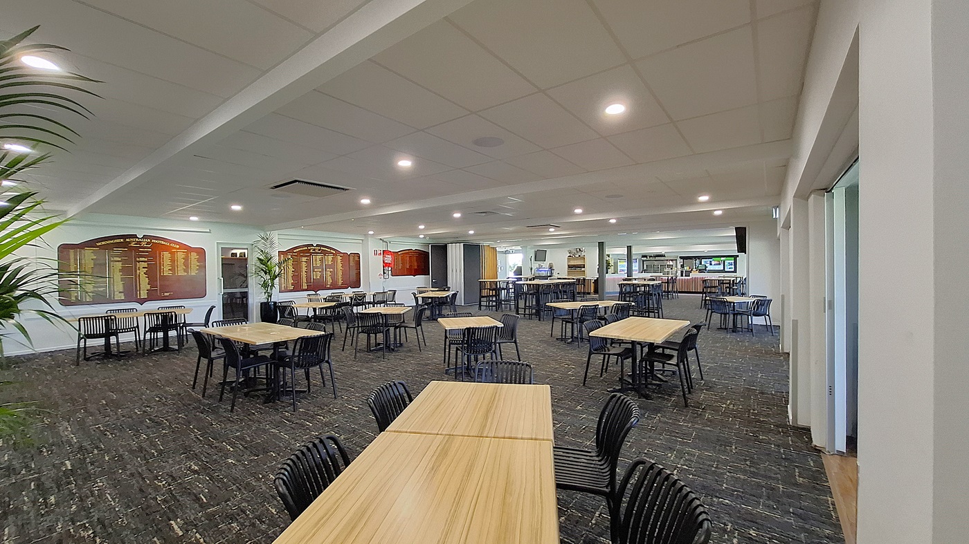 On the left, a large potted plant adds a touch of greenery beside a wall featuring framed boards with text. Several wooden tables with black chairs are arranged across the dark carpeted floor. In the centre, the space is open, with more tables visible, creating a dining area. To the right, a bar counter is seen in the background, with shelves and equipment. The ceiling is white with recessed lighting, providing a bright atmosphere throughout the room. On the left, a large potted plant adds a touch of greenery beside a wall featuring framed boards with text. Several wooden tables with black chairs are arranged across the dark carpeted floor. In the centre, the space is open, with more tables visible, creating a dining area. To the right, a bar counter is seen in the background, with shelves and equipment. The ceiling is white with recessed lighting, providing a bright atmosphere throughout the room.