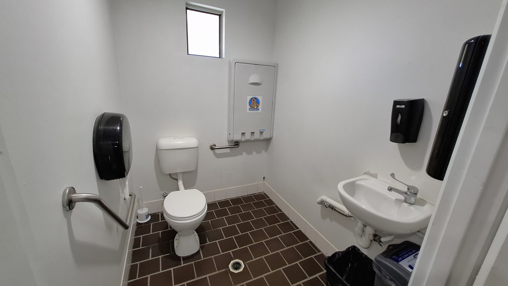 On the left, a white toilet is positioned next to a wall-mounted paper towel dispenser. A grab bar is installed beside the toilet for support. In the centre, the floor is covered with dark brown tiles arranged in a grid pattern, leading to a drain in the middle. To the right, a small sink is mounted on the wall, with a tap and a waste bin underneath. A small window above the toilet allows light into the space. The walls are painted white, and the ceiling is also white. On the left, a white toilet is positioned next to a wall-mounted paper towel dispenser. A grab bar is installed beside the toilet for support. In the centre, the floor is covered with dark brown tiles arranged in a grid pattern, leading to a drain in the middle. To the right, a small sink is mounted on the wall, with a tap and a waste bin underneath. A small window above the toilet allows light into the space. The walls are painted white, and the ceiling is also white.