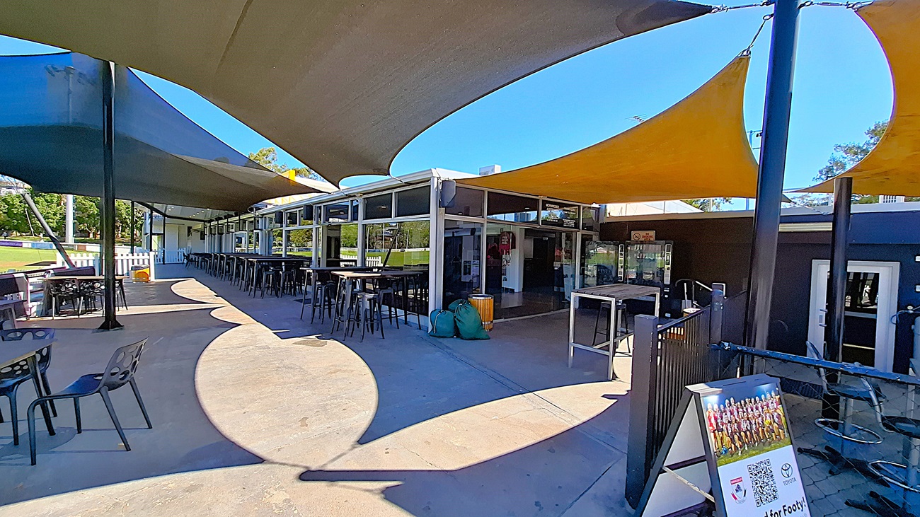 On the left, a wooden bin is positioned next to a green bag, while a glass door leads into the club. To the right, two vending machines are visible, displaying various snacks and drinks. In the centre, the entrance opens to a spacious area with wooden flooring, leading to tall tables and chairs arranged throughout. The ceiling is covered with a tan awning, providing shade. Above the entrance, a sign displays the club's name and trading hours. On the left, a wooden bin is positioned next to a green bag, while a glass door leads into the club. To the right, two vending machines are visible, displaying various snacks and drinks. In the centre, the entrance opens to a spacious area with wooden flooring, leading to tall tables and chairs arranged throughout. The ceiling is covered with a tan awning, providing shade. Above the entrance, a sign displays the club's name and trading hours.