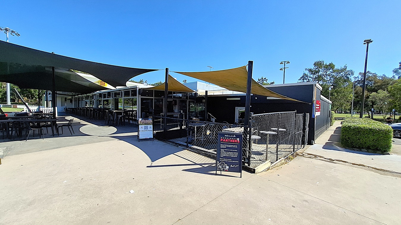 On the left, a series of black tables and chairs are arranged under a large green shade sail, providing outdoor seating. The floor is made of smooth concrete, leading towards a fenced area. In the centre, the entrance to the building features large glass windows, allowing visibility into the interior. To the right, a sign stands on the ground, displaying information about the venue. The ceiling is formed by the shade sails, which are yellow and black, On the left, a series of black tables and chairs are arranged under a large green shade sail, providing outdoor seating. The floor is made of smooth concrete, leading towards a fenced area. In the centre, the entrance to the building features large glass windows, allowing visibility into the interior. To the right, a sign stands on the ground, displaying information about the venue. The ceiling is formed by the shade sails, which are yellow and black,