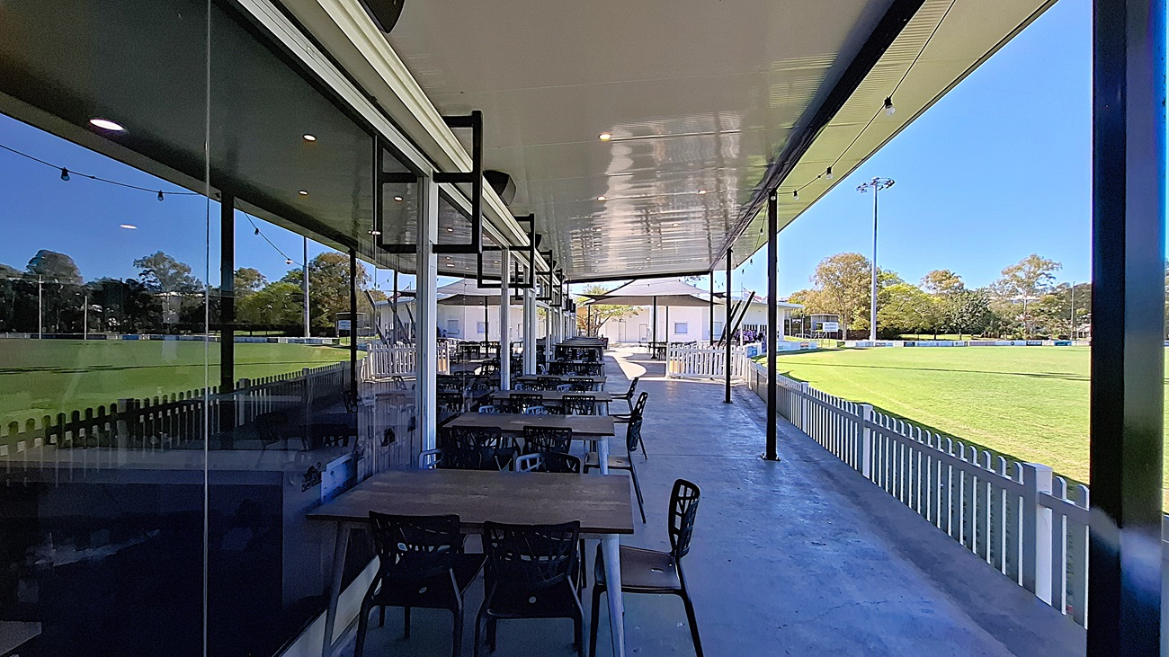 On the left, a row of wooden tables with black chairs is positioned under a covered area, with large glass windows reflecting the surroundings. The floor is smooth and grey, extending towards the right. In the background, a green field is visible, bordered by a white picket fence. To the right, additional seating is arranged, leading to a building with white walls. The ceiling is flat and white, with overhead lights providing illumination. On the left, a row of wooden tables with black chairs is positioned under a covered area, with large glass windows reflecting the surroundings. The floor is smooth and grey, extending towards the right. In the background, a green field is visible, bordered by a white picket fence. To the right, additional seating is arranged, leading to a building with white walls. The ceiling is flat and white, with overhead lights providing illumination.