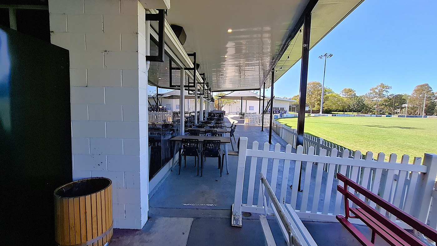 On the left, a green wall is visible next to a wooden bin, while a series of tables and chairs are arranged along the covered area. The floor is smooth and grey, extending towards the right. In the background, a white picket fence borders a green field, with several buildings visible beyond it. To the right, a red bench is positioned against the fence. The ceiling is flat and white, with overhead lights. On the left, a green wall is visible next to a wooden bin, while a series of tables and chairs are arranged along the covered area. The floor is smooth and grey, extending towards the right. In the background, a white picket fence borders a green field, with several buildings visible beyond it. To the right, a red bench is positioned against the fence. The ceiling is flat and white, with overhead lights.