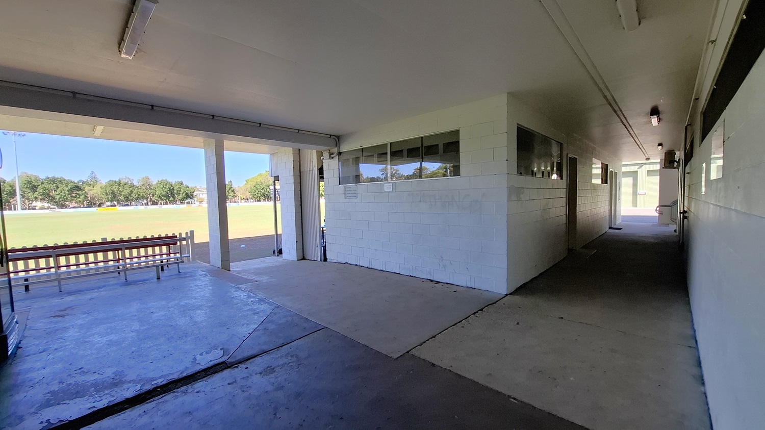 On the left, a concrete wall features a series of windows, allowing light into the corridor. A bench with a red seat is positioned against the wall, facing an open area. The floor is a mix of concrete and tiles, leading towards a grassy field visible in the background. To the right, a narrow corridor extends, with doors along the wall and a light fixture above. The ceiling is flat and white, with fluorescent lights providing illumination throughout the space. On the left, a concrete wall features a series of windows, allowing light into the corridor. A bench with a red seat is positioned against the wall, facing an open area. The floor is a mix of concrete and tiles, leading towards a grassy field visible in the background. To the right, a narrow corridor extends, with doors along the wall and a light fixture above. The ceiling is flat and white, with fluorescent lights providing illumination throughout the space.