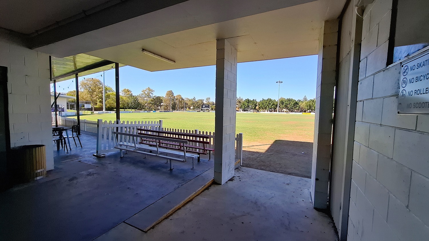 On the left, a wooden bin is positioned next to a wall, with a small table and chairs visible nearby. The floor is a combination of concrete and tiles, leading to an open area. In the foreground, a white picket fence separates the space from a grassy field, which extends into the background. To the right, a wall features a sign with various rules. The ceiling is flat and white, with overhead lights. On the left, a wooden bin is positioned next to a wall, with a small table and chairs visible nearby. The floor is a combination of concrete and tiles, leading to an open area. In the foreground, a white picket fence separates the space from a grassy field, which extends into the background. To the right, a wall features a sign with various rules. The ceiling is flat and white, with overhead lights.