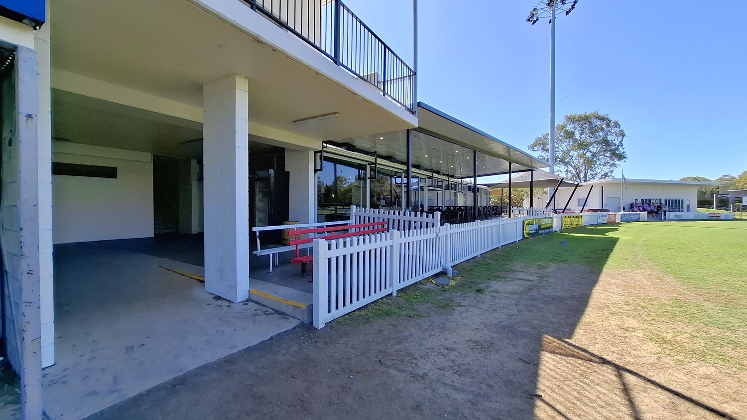 On the left, a concrete area leads to a covered space with a white railing and a red bench. The floor is a mix of concrete and grass, transitioning to a grassy field in the foreground. In the background, a large glass structure features multiple windows, providing views of the interior seating. To the right, another building is visible, with a similar design. The ceiling above the covered area is flat and extends outwards, providing shelter while allowing light to enter through the glass panels. On the left, a concrete area leads to a covered space with a white railing and a red bench. The floor is a mix of concrete and grass, transitioning to a grassy field in the foreground. In the background, a large glass structure features multiple windows, providing views of the interior seating. To the right, another building is visible, with a similar design. The ceiling above the covered area is flat and extends outwards, providing shelter while allowing light to enter through the glass panels.