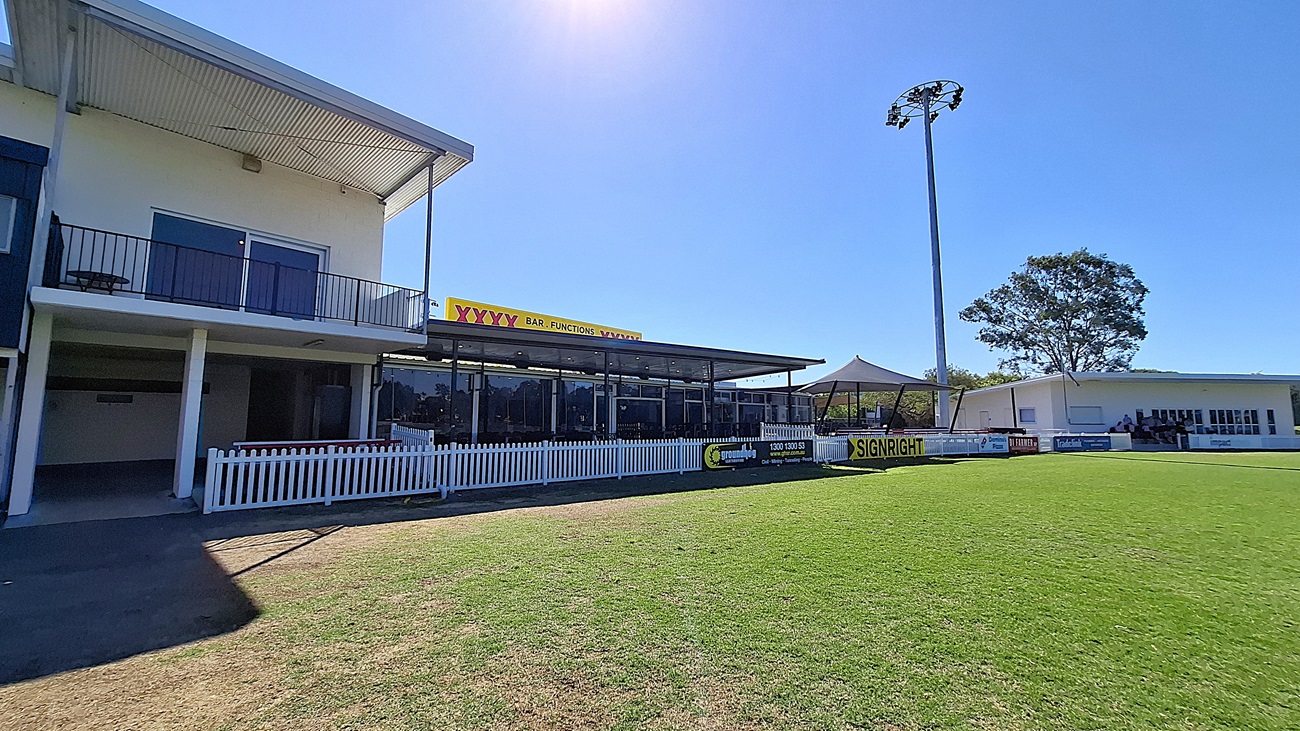 On the left, a two-storey building features a balcony with black railings and large windows. Below, a covered area with glass panels provides seating. The floor is a mix of grass and concrete, leading to a well-maintained green field in the foreground. To the right, another building with white walls and large windows is visible, along with a tall light pole. The ceiling of the covered area is flat and extends outwards. A white picket fence runs along the front. On the left, a two-storey building features a balcony with black railings and large windows. Below, a covered area with glass panels provides seating. The floor is a mix of grass and concrete, leading to a well-maintained green field in the foreground. To the right, another building with white walls and large windows is visible, along with a tall light pole. The ceiling of the covered area is flat and extends outwards. A white picket fence runs along the front.