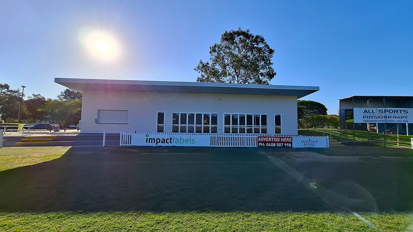 On the left, a modern building features large glass windows and a white sign with "impact labels" displayed prominently. Steps lead up to the entrance, which is flanked by a grassy area. The floor is a mix of grass and concrete, extending towards the right. To the right, another building is visible with a sign for "All Sports Physiotherapy." The ceiling of the main building is flat and white, providing a clean, functional appearance. On the left, a modern building features large glass windows and a white sign with "impact labels" displayed prominently. Steps lead up to the entrance, which is flanked by a grassy area. The floor is a mix of grass and concrete, extending towards the right. To the right, another building is visible with a sign for "All Sports Physiotherapy." The ceiling of the main building is flat and white, providing a clean, functional appearance.