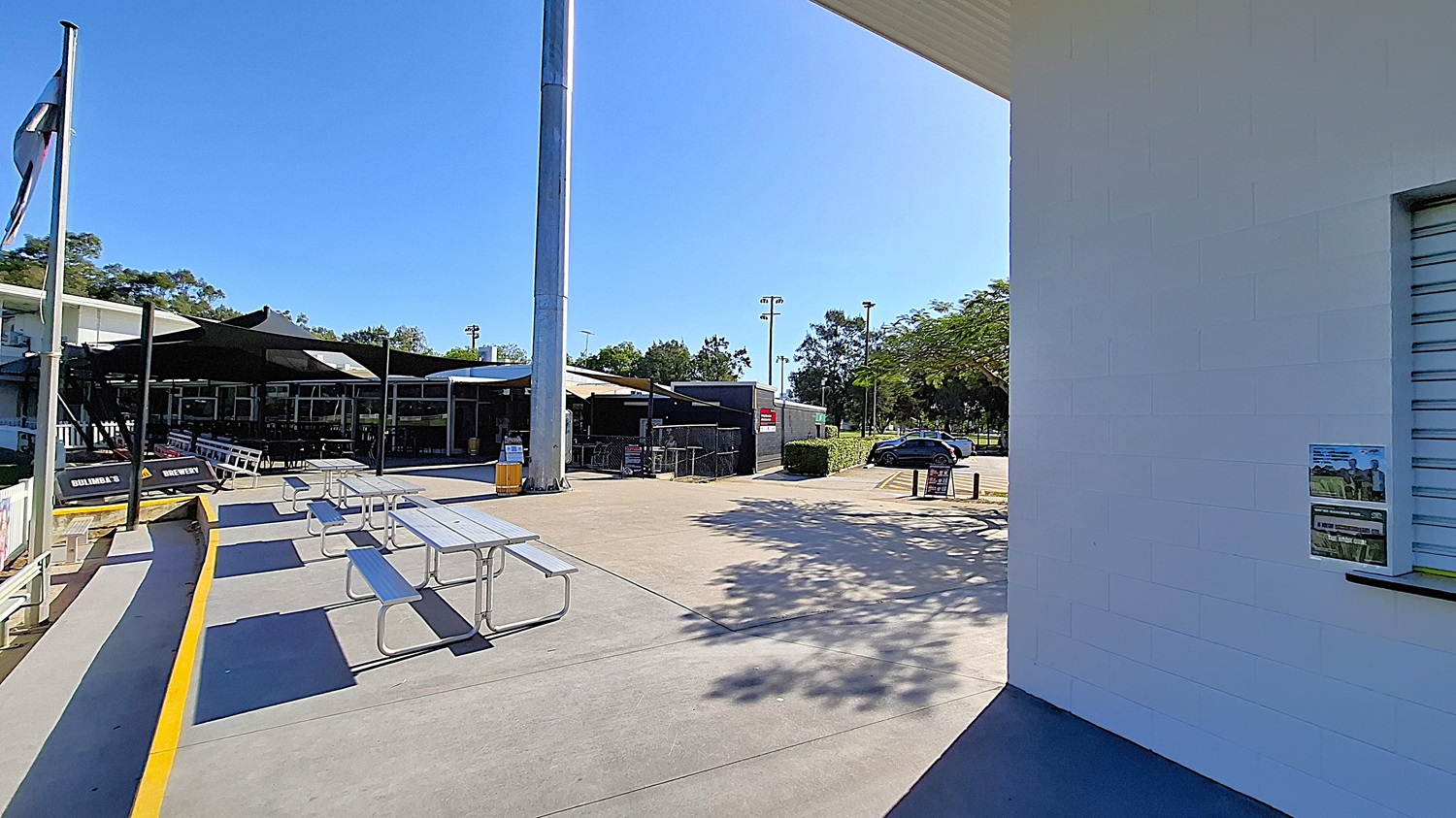 On the left, a series of picnic tables are arranged on a concrete surface, with a yellow edge marking the area. A tall light pole stands nearby, and a shaded seating area is visible in the background, featuring large glass windows. The floor is smooth concrete, extending towards a grassy area in the foreground. To the right, a white wall with a small window displays a poster. The ceiling above the seating area is flat and extends outwards. On the left, a series of picnic tables are arranged on a concrete surface, with a yellow edge marking the area. A tall light pole stands nearby, and a shaded seating area is visible in the background, featuring large glass windows. The floor is smooth concrete, extending towards a grassy area in the foreground. To the right, a white wall with a small window displays a poster. The ceiling above the seating area is flat and extends outwards.