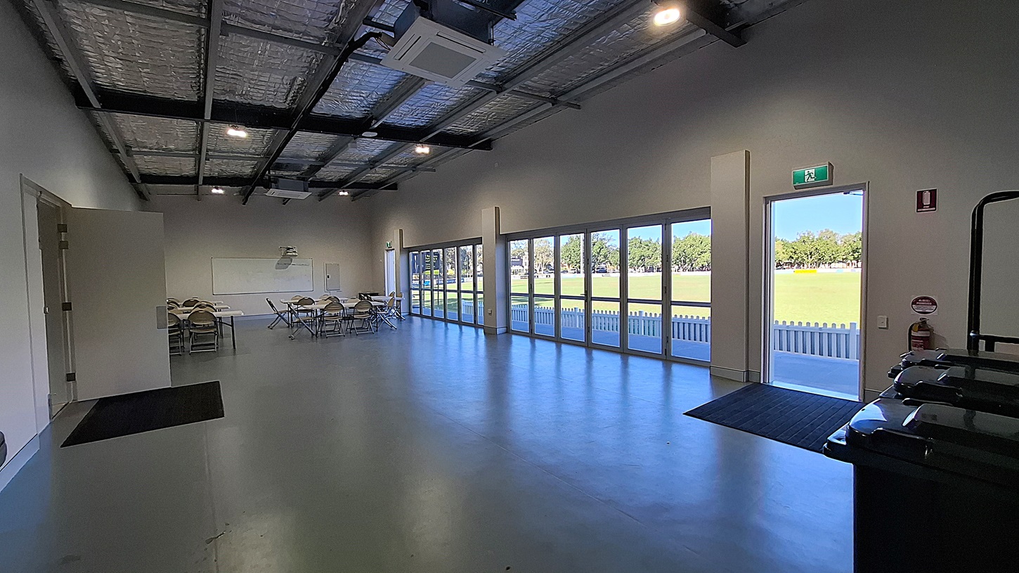On the left, a small meeting area features several folding chairs and tables arranged neatly. A whiteboard is mounted on the wall, and a door leads to another room. The floor is smooth and grey, extending towards the right side. To the right, large glass doors open to a grassy area outside, with a white picket fence visible beyond. The ceiling is high with exposed beams and insulation, fitted with overhead lights. An exit sign is mounted above the door, indicating the way out. On the left, a small meeting area features several folding chairs and tables arranged neatly. A whiteboard is mounted on the wall, and a door leads to another room. The floor is smooth and grey, extending towards the right side. To the right, large glass doors open to a grassy area outside, with a white picket fence visible beyond. The ceiling is high with exposed beams and insulation, fitted with overhead lights. An exit sign is mounted above the door, indicating the way out.