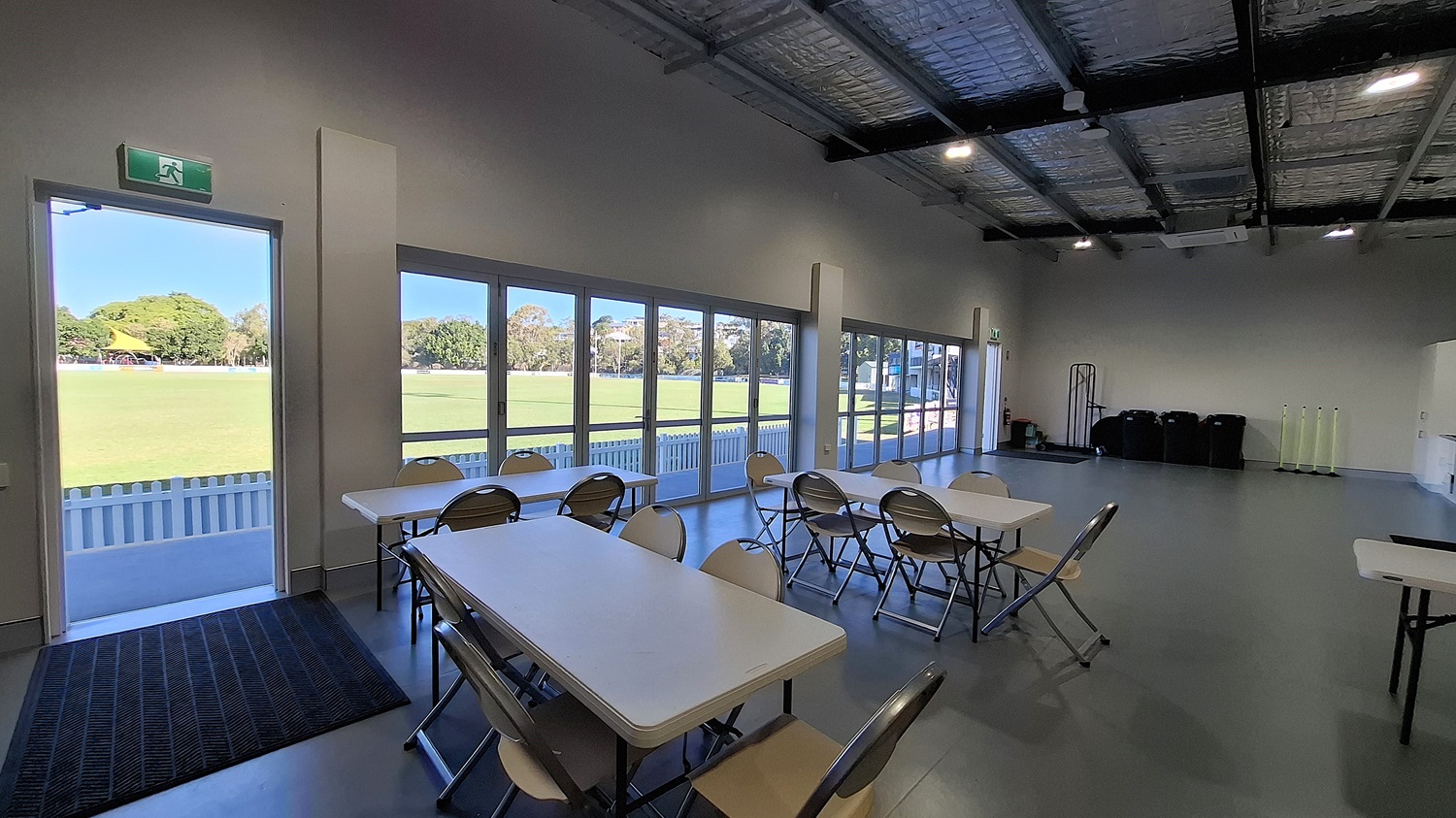 On the left, a doorway leads outside, framed by a grey wall. A black mat is positioned at the entrance, and large glass doors provide a view of a grassy field beyond. Several white tables and folding chairs are arranged in the middle of the room, with a smooth grey floor beneath. To the right, a wall features a small window and a few bins stacked against it. The ceiling is high with exposed beams and insulation, fitted with overhead lights . On the left, a doorway leads outside, framed by a grey wall. A black mat is positioned at the entrance, and large glass doors provide a view of a grassy field beyond. Several white tables and folding chairs are arranged in the middle of the room, with a smooth grey floor beneath. To the right, a wall features a small window and a few bins stacked against it. The ceiling is high with exposed beams and insulation, fitted with overhead lights .