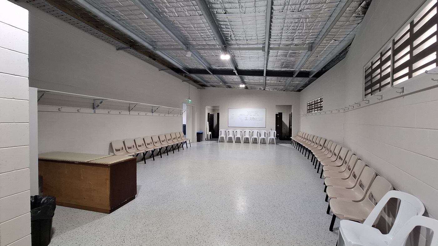 On the left, a wooden table is positioned against the wall, with a black bin nearby. Above, a row of hooks is mounted for hanging items. The floor is smooth and light-coloured, extending throughout the room. In the middle, a whiteboard is mounted on the wall, with several chairs arranged in a semi-circle around the space. To the right, more chairs are lined up against the wall, and a doorway leads to another area. The ceiling is flat with exposed beams and insulation, fitted with overhead lights On the left, a wooden table is positioned against the wall, with a black bin nearby. Above, a row of hooks is mounted for hanging items. The floor is smooth and light-coloured, extending throughout the room. In the middle, a whiteboard is mounted on the wall, with several chairs arranged in a semi-circle around the space. To the right, more chairs are lined up against the wall, and a doorway leads to another area. The ceiling is flat with exposed beams and insulation, fitted with overhead lights