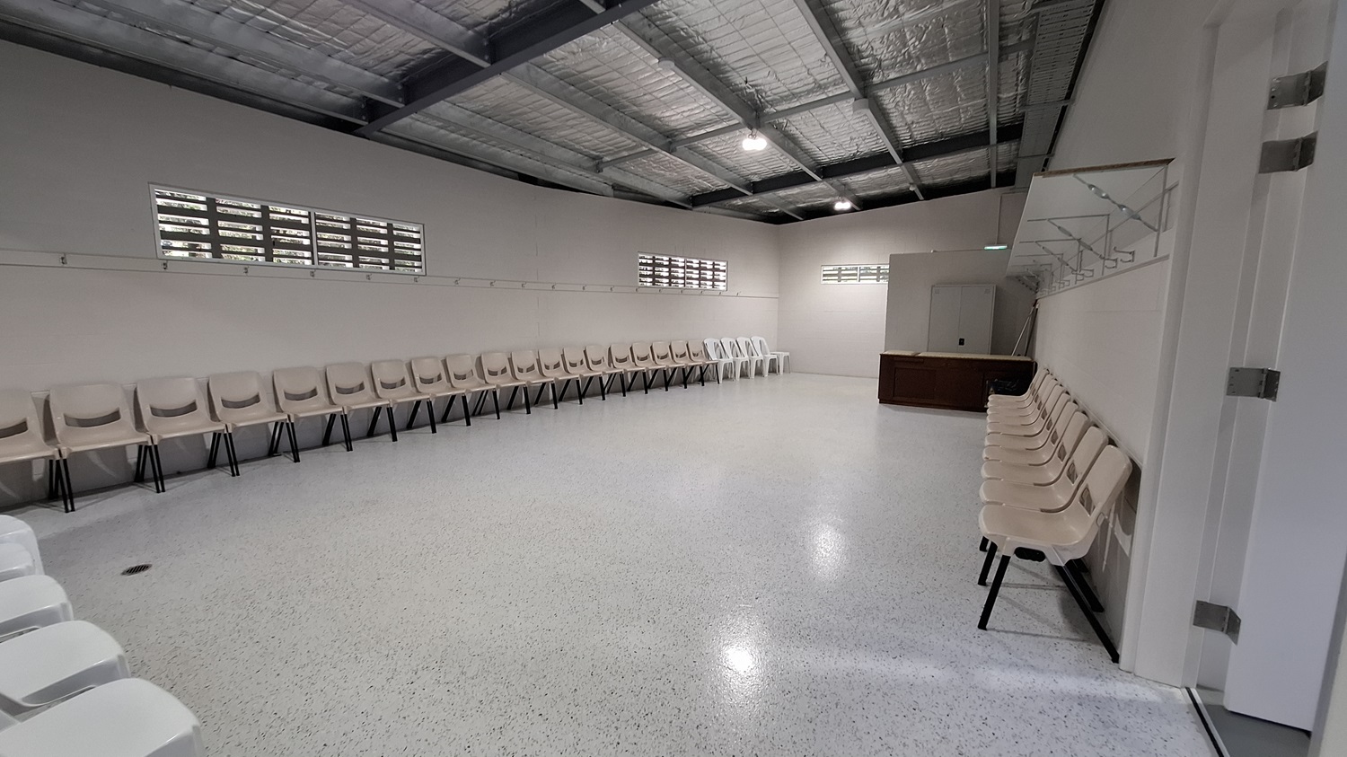 On the left, a row of beige chairs is arranged along the wall, with a few white chairs interspersed. Above, small windows allow light to enter, and a rail runs along the wall for hanging items. The floor is smooth and polished, reflecting the light. In the middle, the space is open, leading to a wooden counter against the right wall. The ceiling is high with exposed beams and insulation, fitted with overhead lights. On the left, a row of beige chairs is arranged along the wall, with a few white chairs interspersed. Above, small windows allow light to enter, and a rail runs along the wall for hanging items. The floor is smooth and polished, reflecting the light. In the middle, the space is open, leading to a wooden counter against the right wall. The ceiling is high with exposed beams and insulation, fitted with overhead lights.