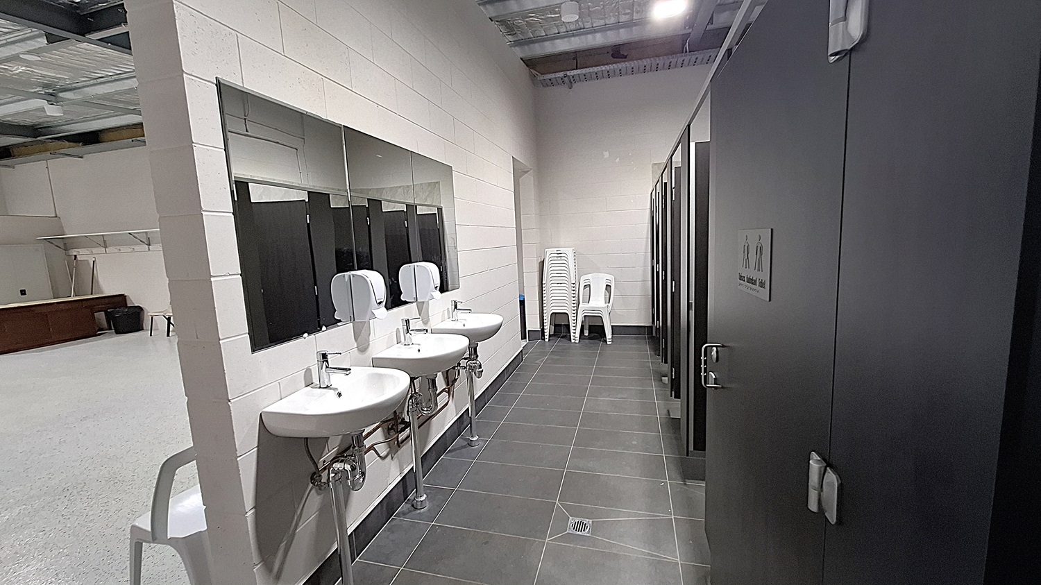 On the left, three white sinks are mounted on the wall, each with a soap dispenser above. A large mirror spans the length of the sinks. The floor is dark grey tiles, extending towards the right. To the right, several black cubicle doors are visible, each with a sign indicating male and female facilities. A stack of white plastic chairs is positioned against the wall. The ceiling is high with exposed metal beams and panels, fitted with overhead lights. On the left, three white sinks are mounted on the wall, each with a soap dispenser above. A large mirror spans the length of the sinks. The floor is dark grey tiles, extending towards the right. To the right, several black cubicle doors are visible, each with a sign indicating male and female facilities. A stack of white plastic chairs is positioned against the wall. The ceiling is high with exposed metal beams and panels, fitted with overhead lights.