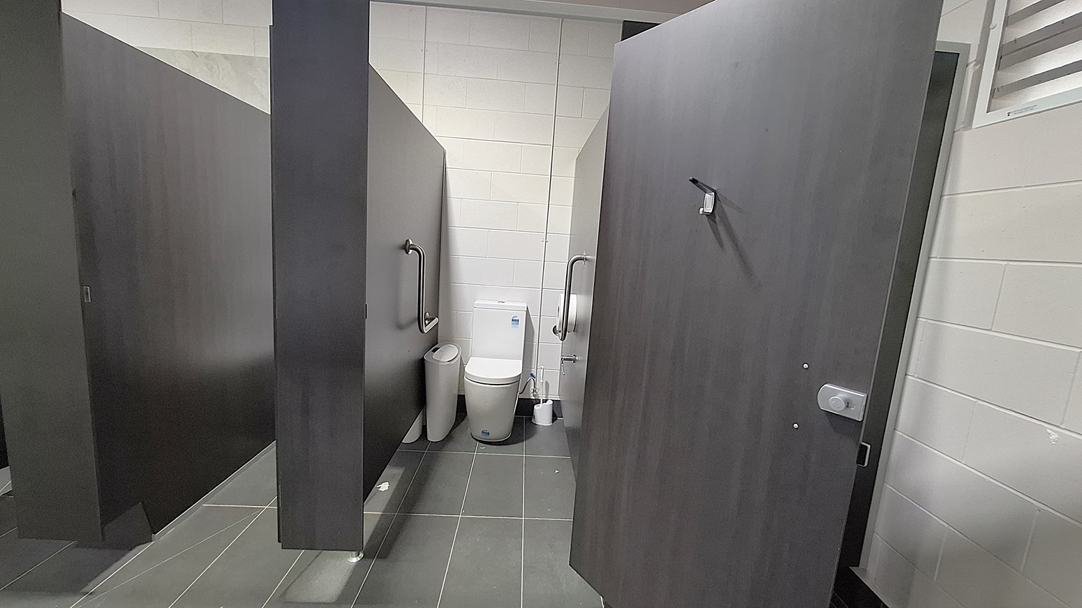 On the left, a dark wooden cubicle door stands closed, with a grab rail mounted beside it. Adjacent to it, a white toilet is positioned against the wall, with a small bin nearby. The floor is dark grey tiles, extending throughout the space. To the right, another cubicle door is slightly ajar, revealing a similar setup. The ceiling is flat and white, with ventilation grilles visible, providing a clean and functional environment. On the left, a dark wooden cubicle door stands closed, with a grab rail mounted beside it. Adjacent to it, a white toilet is positioned against the wall, with a small bin nearby. The floor is dark grey tiles, extending throughout the space. To the right, another cubicle door is slightly ajar, revealing a similar setup. The ceiling is flat and white, with ventilation grilles visible, providing a clean and functional environment.