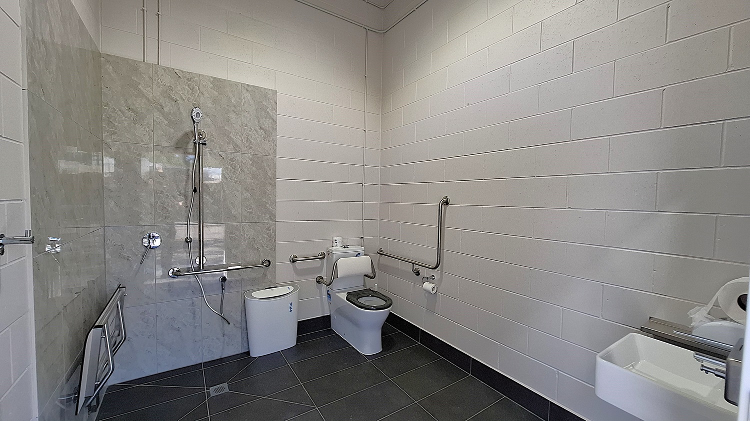 On the left, a shower area features a wall of grey tiles with a handheld showerhead mounted above. A grab rail is installed beside the shower. Adjacent to it, a white toilet is positioned with a roll of toilet paper mounted on the wall. The floor is dark grey tiles, extending throughout the space. To the right, a small sink is mounted on the wall, with a paper towel dispenser nearby. The ceiling is flat and white, with exposed pipes running along it. On the left, a shower area features a wall of grey tiles with a handheld showerhead mounted above. A grab rail is installed beside the shower. Adjacent to it, a white toilet is positioned with a roll of toilet paper mounted on the wall. The floor is dark grey tiles, extending throughout the space. To the right, a small sink is mounted on the wall, with a paper towel dispenser nearby. The ceiling is flat and white, with exposed pipes running along it.