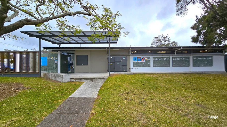 Grey and white cemented pathway leading to the facility entrance. There is a large open space in the front of the facility that is covered with grass. A grey metal locker cabinet is placed against the right wall, which is sun-protected by a skylight roofing supported by a metal roof frame. A few meters away are four windows with some small adverts in the first window frame. Grey and white cemented pathway leading to the facility entrance. There is a large open space in the front of the facility that is covered with grass. A grey metal locker cabinet is placed against the right wall, which is sun-protected by a skylight roofing supported by a metal roof frame. A few meters away are four windows with some small adverts in the first window frame.